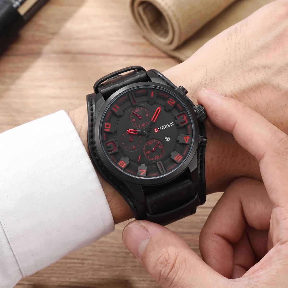 Man wearing a black watch with red numerals and a wide leather cuff strap with a white shirt cuff