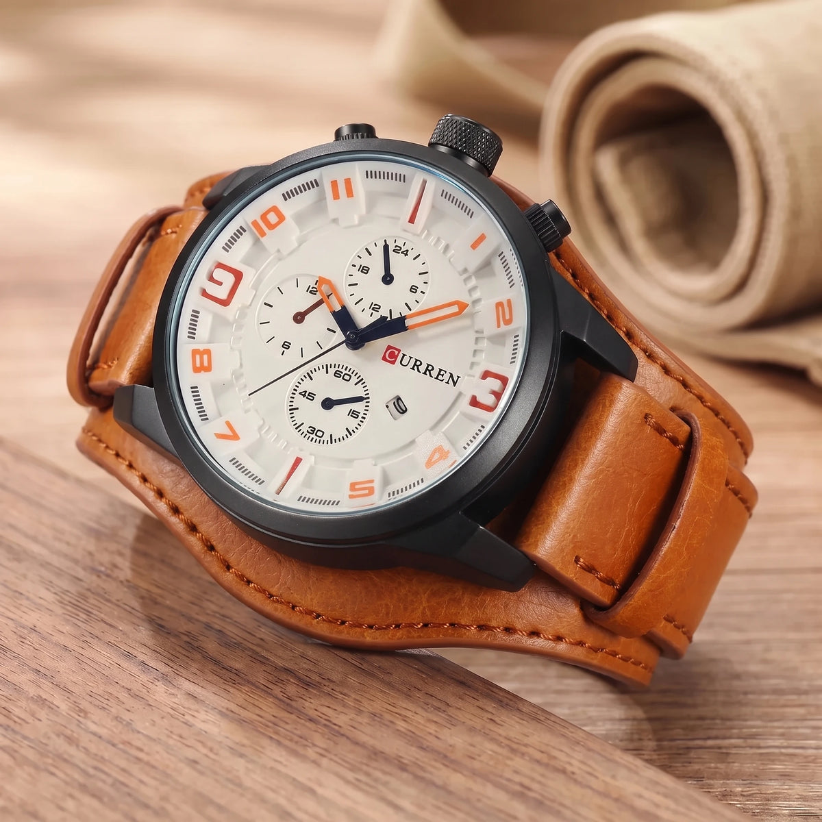 White dial watch with orange numerals and a wide tan leather cuff strap on a walnut table