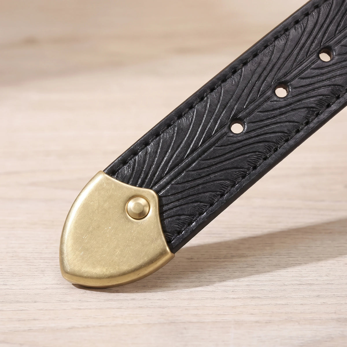 Close-up of the brass shield tip and feather pattern on the black leather belt strap